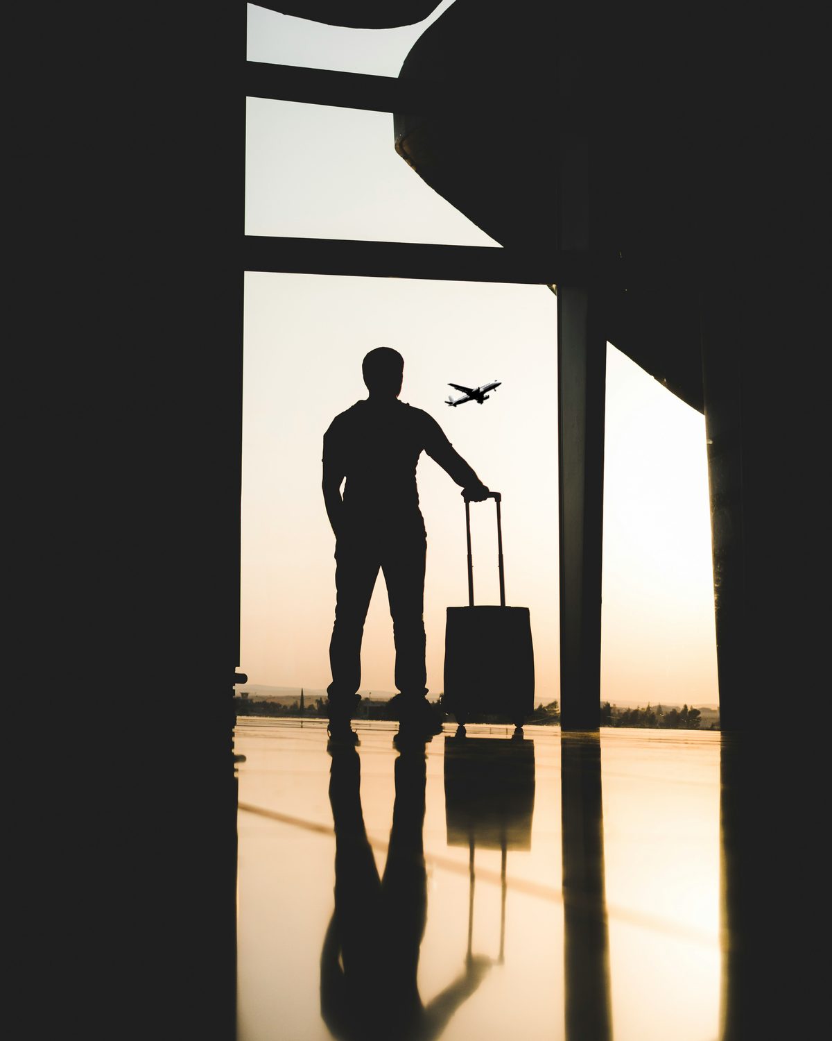A traveller watching a plane take off from an airport window at sunset